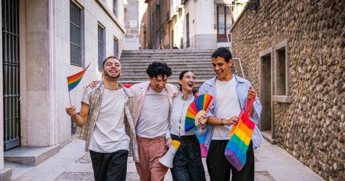 Four happy diverse friends walking together in a city street, carrying rainbow flags and a fan, celebrating lgbtq plus pride