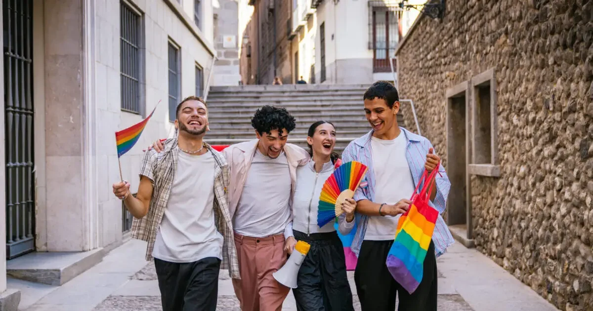 Four happy diverse friends walking together in a city street, carrying rainbow flags and a fan, celebrating lgbtq plus pride