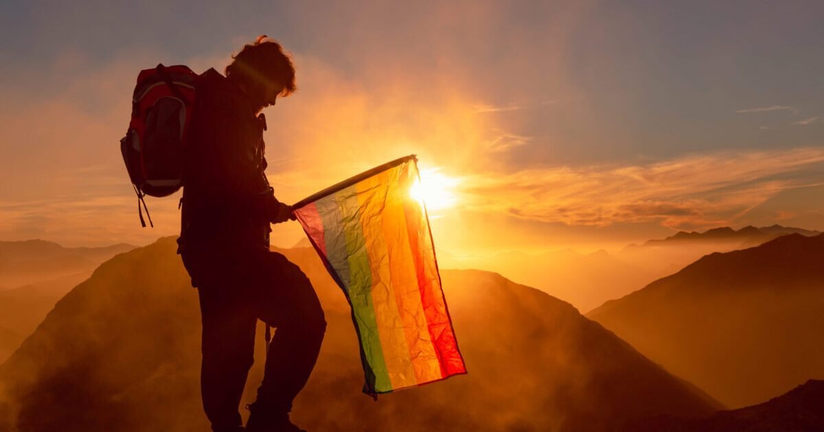 backlit silhouette of hiker with backpack carrying a rainbow LGBT pride flag on top of a mountain watching the sunset. sport and healthy life concept.